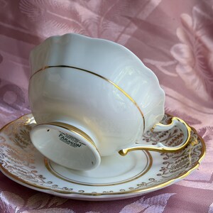 May include: A white porcelain teacup and saucer set featuring a scalloped rim and gold trim. The teacup base is marked with the text PARAGON. The saucer and cup handle are decorated with intricate gold filigree patterns against the white surface.