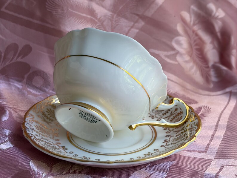 May include: A white porcelain teacup and saucer set featuring a scalloped rim and gold trim. The teacup base is marked with the text PARAGON. The saucer and cup handle are decorated with intricate gold filigree patterns against the white surface.