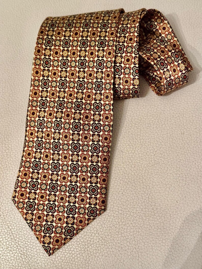 May include: A necktie featuring a repeating geometric pattern of small, stylized four-petaled floral shapes in shades of dark brown, rust orange, and cream. The fabric has a smooth, slightly reflective finish, and the tie is folded at the top.