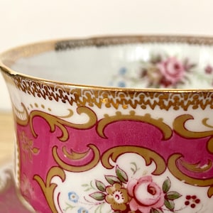 May include: Close-up of a vintage teacup with a vibrant pink and gold design. The cup features ornate gold detailing, a scalloped rim, and a floral pattern with pink roses and blue accents. The teacup is a classic piece of tableware.