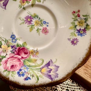 May include: A white porcelain saucer with a gold rim and a floral design. The saucer features a variety of flowers in shades of pink, purple, yellow, and blue.
