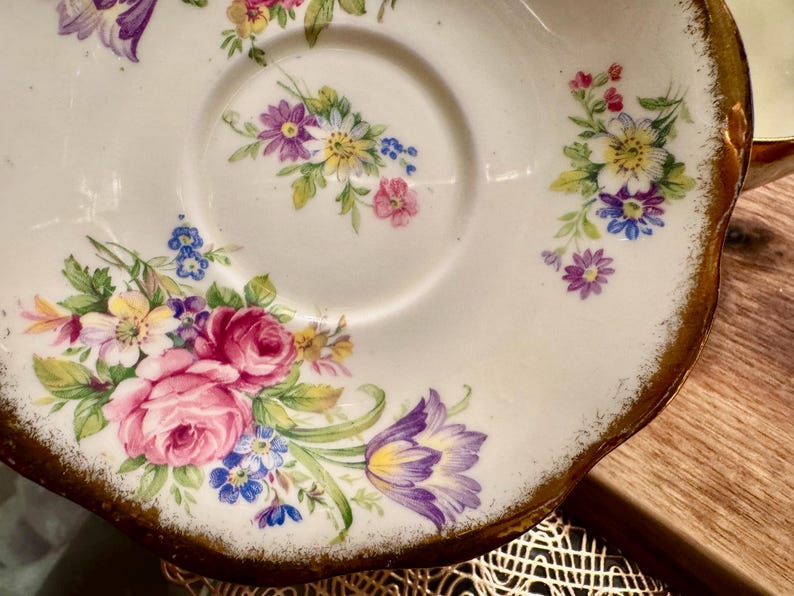 May include: A white porcelain saucer with a gold rim and a floral design. The saucer features a variety of flowers in shades of pink, purple, yellow, and blue.