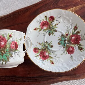 May include: A white teacup and saucer set with a floral design featuring red roses and green leaves. The set has a gold trim around the edge of the saucer.