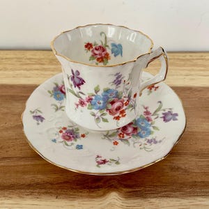 May include: A white teacup and saucer set with a floral pattern in pink, blue, and purple. The cup and saucer have a scalloped edge and gold trim. The teacup sits on the saucer, ready for tea.
