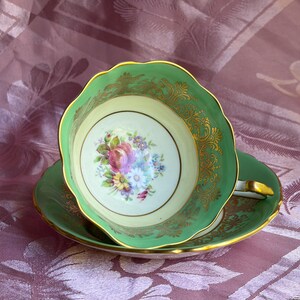 Peut inclure: Un ensemble tasse et soucoupe en porcelaine avec un bord vert p&acirc;le orn&eacute; de d&eacute;tails en filigrane dor&eacute;. L'int&eacute;rieur de la tasse pr&eacute;sente un bouquet floral central compos&eacute; de fleurs roses, violettes et jaunes sur un fond cr&egrave;me.
