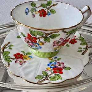 K&ouml;nnte beinhalten: Wei&szlig;es Tee-Set mit einem Blumendesign aus roten, blauen und gelben Bl&uuml;ten auf braunen Zweigen mit gr&uuml;nen Bl&auml;ttern. Tasse und Untertasse haben einen Goldrand. Die Untertasse hat einen gewellten Rand und einen gr&uuml;nen Sockel.