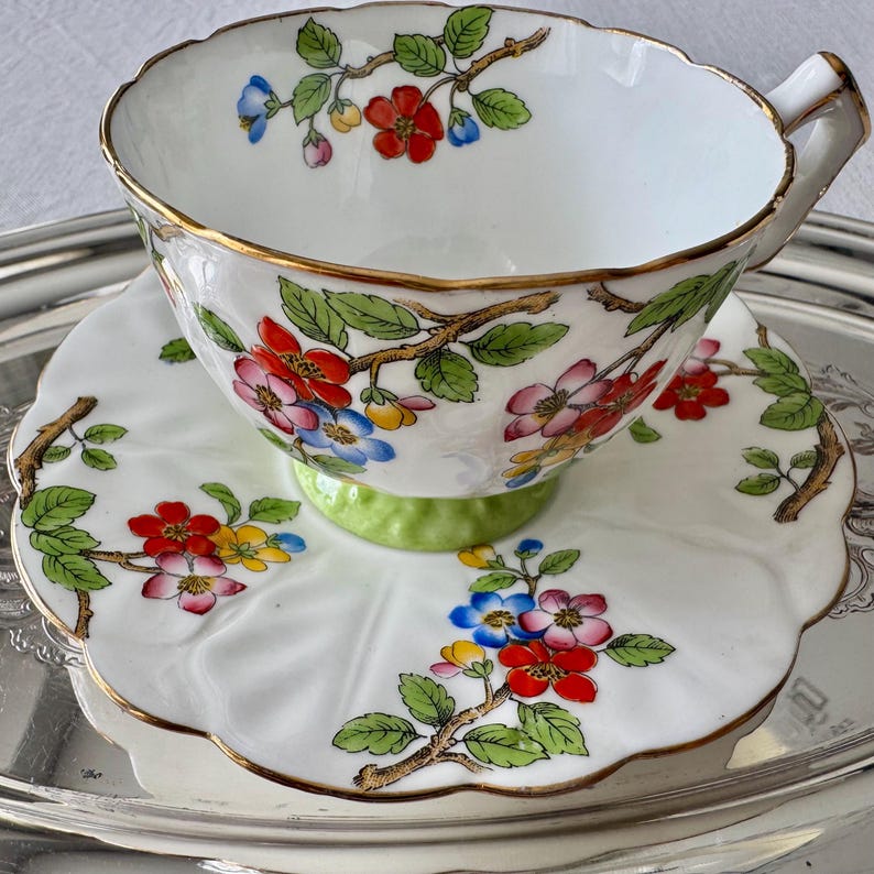 K&ouml;nnte beinhalten: Wei&szlig;es Tee-Set mit einem Blumendesign aus roten, blauen und gelben Bl&uuml;ten auf braunen Zweigen mit gr&uuml;nen Bl&auml;ttern. Tasse und Untertasse haben einen Goldrand. Die Untertasse hat einen gewellten Rand und einen gr&uuml;nen Sockel.