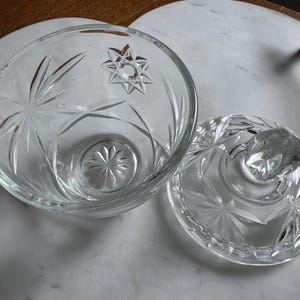 May include: A clear glass bowl with a matching lid, featuring a pressed starburst and geometric pattern. The bowl has a circular base with a radiating floral design, and the lid includes a faceted knob handle for easy removal and placement.