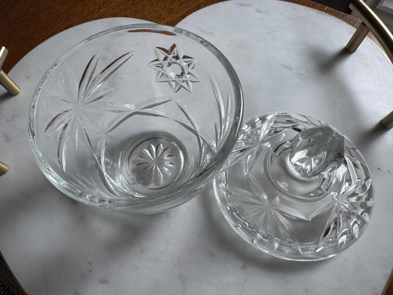 May include: A clear glass bowl with a matching lid, featuring a pressed starburst and geometric pattern. The bowl has a circular base with a radiating floral design, and the lid includes a faceted knob handle for easy removal and placement.