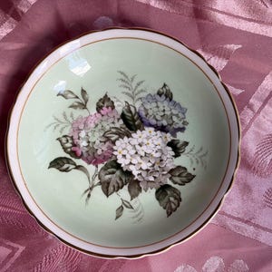 May include: A round ceramic bowl with a pale mint green interior featuring a central floral illustration of pink, white, and blue hydrangea blossoms with dark green leaves. The bowl has a scalloped edge trimmed with a thin gold metallic band.