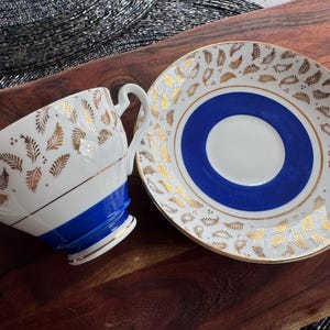 May include: A white teacup and saucer with a blue and gold floral pattern. The teacup has a blue band around the top and the saucer has a blue center circle.