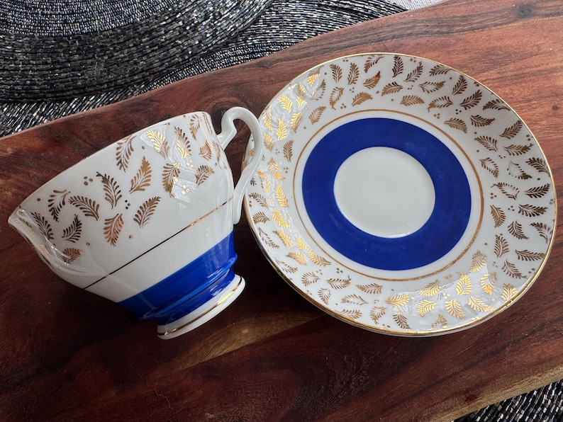 May include: A white teacup and saucer with a blue and gold floral pattern. The teacup has a blue band around the top and the saucer has a blue center circle.