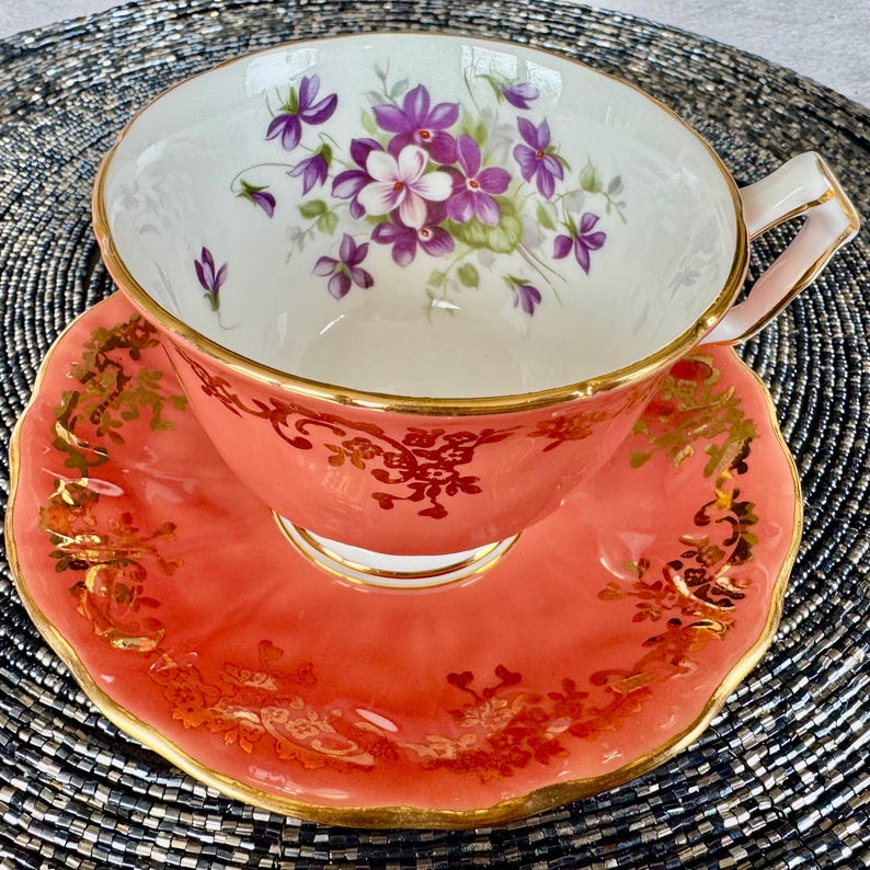 May include: A vintage teacup and saucer set with a delicate floral design. The cup and saucer are both a vibrant coral color with gold accents. The floral design features purple and white violets.