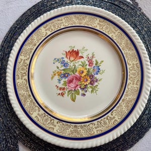 May include: A vintage porcelain plate with a floral bouquet centerpiece featuring a red tulip, pink rose, and other colorful flowers. The plate has a cream-colored border with gold and blue accents, and a white, rope-like edge.