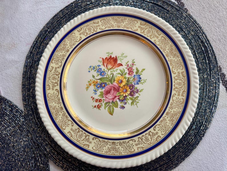 May include: A vintage porcelain plate with a floral bouquet centerpiece featuring a red tulip, pink rose, and other colorful flowers. The plate has a cream-colored border with gold and blue accents, and a white, rope-like edge.