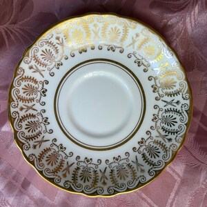 May include: A white porcelain saucer featuring a scalloped edge and a gold-toned rim. The border is decorated with a repeating pattern of gold filigree, stylized floral motifs, and fan-shaped elements surrounding a plain white center.
