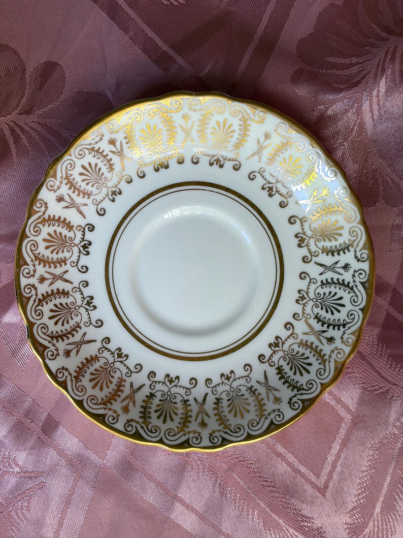 May include: A white porcelain saucer featuring a scalloped edge and a gold-toned rim. The border is decorated with a repeating pattern of gold filigree, stylized floral motifs, and fan-shaped elements surrounding a plain white center.