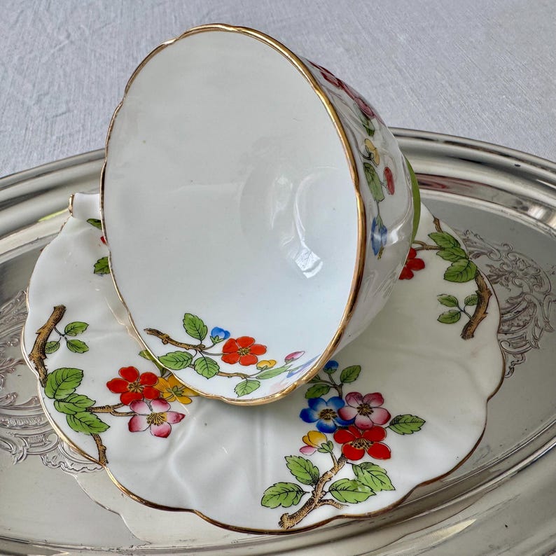 K&ouml;nnte beinhalten: Wei&szlig;es Tee-Set mit Blumendesign. Die Tasse und Untertasse haben einen Goldrand und sind mit roten, blauen und gelben Blumen und gr&uuml;nen Bl&auml;ttern verziert. Die Tasse liegt auf der Untertasse, die sich auf einem Silbertablett befindet.