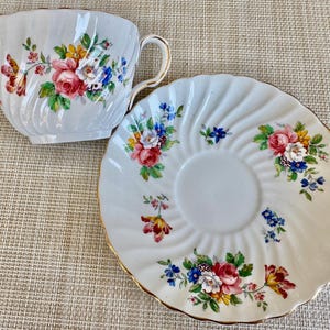 Puede incluir: Una taza de t&eacute; y un platillo blancos con un patr&oacute;n floral. La taza y el platillo tienen un borde dorado. Las flores son rosas, amarillas, azules y verdes.