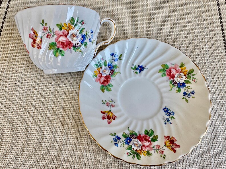 Puede incluir: Una taza de t&eacute; y un platillo blancos con un patr&oacute;n floral. La taza y el platillo tienen un borde dorado. Las flores son rosas, amarillas, azules y verdes.