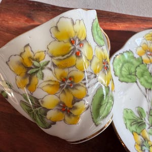 May include: A white teacup and saucer with a floral design. The design features yellow flowers with red centers and green leaves.