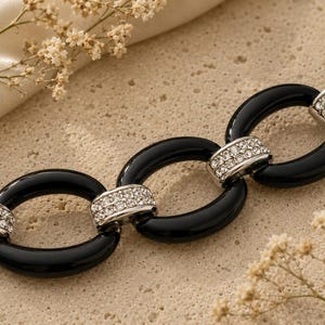 May include: Close-up of a black oval link necklace with silver rhinestone accents. The necklace is displayed on a textured, light-colored surface, with sprigs of dried flowers in the background. Jewelry.