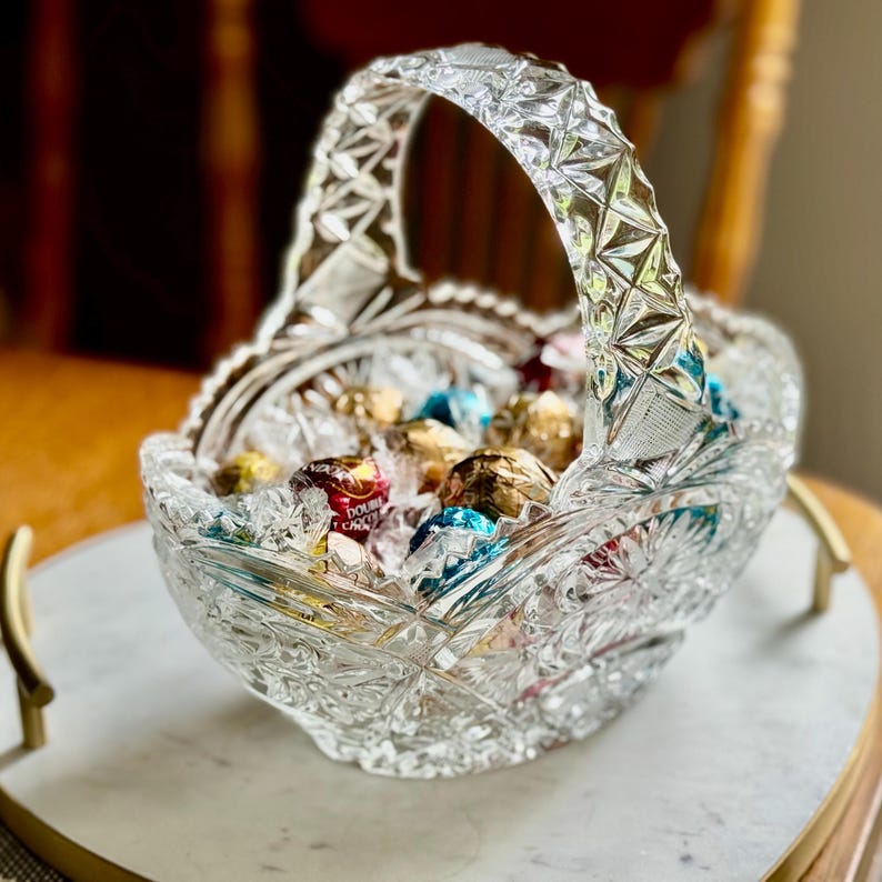 May include: A clear, cut-glass basket with a handle, filled with wrapped chocolates. The basket sits on a white marble tray with gold handles. The glass has a detailed, geometric pattern, reflecting light.