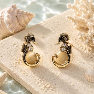 May include: A pair of gold-toned seahorse earrings with black accents and clear crystals. The earrings have a cream-colored pearl-like body. The image is set on a sandy surface with seashells and coral in the background.