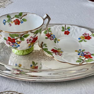 K&ouml;nnte beinhalten: Ein wei&szlig;es Tee-Set mit Blumenmuster in Rot, Blau und Gelb, mit Goldrand. Die Tasse steht auf einem hellgr&uuml;nen Sockel, und das Set steht auf einem silbernen Tablett. Ein klassisches Teeservice.