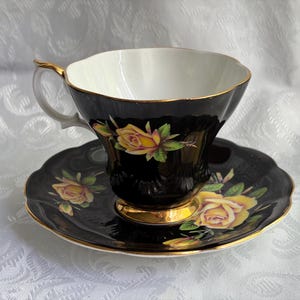 Pode incluir: Um conjunto de ch&aacute;vena e pires em porcelana preta com padr&otilde;es de rosas amarelas e folhas verdes. A ch&aacute;vena tem um interior branco, uma borda recortada e uma asa dourada, enquanto o pires apresenta uma borda dourada recortada a condizer.