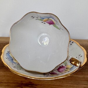 May include: A white teacup and saucer set with a scalloped edge and gold trim. The teacup and saucer feature a floral design with blue, yellow, and pink accents. The teacup is resting on the saucer.