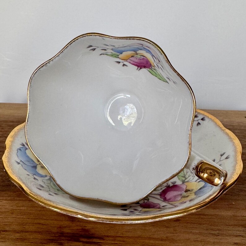 May include: A white teacup and saucer set with a scalloped edge and gold trim. The teacup and saucer feature a floral design with blue, yellow, and pink accents. The teacup is resting on the saucer.