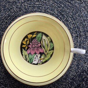 May include: A yellow teacup with a gold rim and a floral design featuring pink, green, and white flowers on a black background.