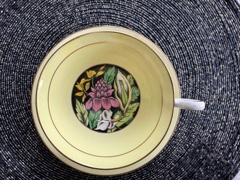 May include: A yellow teacup with a gold rim and a floral design featuring pink, green, and white flowers on a black background.