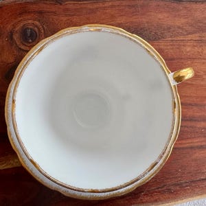 May include: A white teacup with a gold rim and a gold handle. The teacup is sitting on a wooden surface.