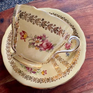 May include: A pale yellow porcelain teacup and saucer set featuring a scalloped edge, a gold leaf border pattern, and a central floral motif with pink, red, and white flowers. The teacup has a curved handle with gold trim and a pedestal base.