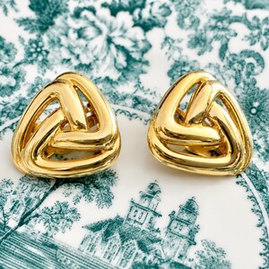 K&ouml;nnte beinhalten: Ein Paar goldfarbene, dreieckige Ohrringe. Jeder Ohrring hat ein gewebtes Design, das eine offene, geometrische Form erzeugt. Die Ohrringe werden auf einem wei&szlig;en Teller mit einem gr&uuml;nen Blumen- und Architekturmuster pr&auml;sentiert.