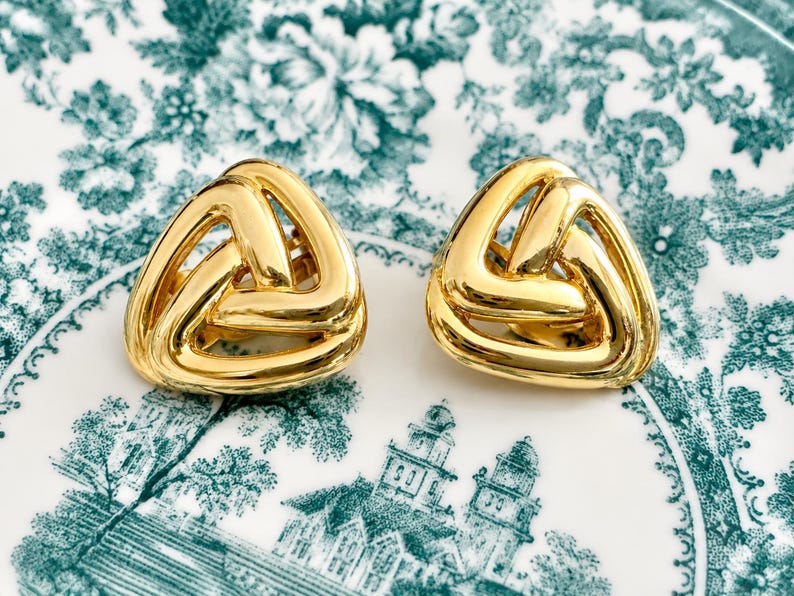 K&ouml;nnte beinhalten: Ein Paar goldfarbene, dreieckige Ohrringe. Jeder Ohrring hat ein gewebtes Design, das eine offene, geometrische Form erzeugt. Die Ohrringe werden auf einem wei&szlig;en Teller mit einem gr&uuml;nen Blumen- und Architekturmuster pr&auml;sentiert.