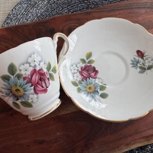 May include: A white teacup and saucer set with a floral design featuring pink and blue flowers. The set has a gold trim around the edges.