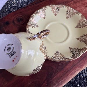 May include: A vintage teacup and saucer set with a pale yellow background and gold floral pattern. The teacup has a gold rim and the saucer has a scalloped edge. The bottom of the teacup has the words "Royal Albert, Made in England, 426" printed in black.
