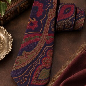Pode incluir: Uma gravata com base azul-escura e um padr&atilde;o paisley complexo em tons de vermelho profundo, verde-azeitona e dourado. O tecido apresenta motivos florais e de gota intrincados dispostos num design sim&eacute;trico ao longo de toda a gravata.