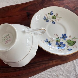 May include: A white teacup and saucer set with a floral design featuring blue flowers and green leaves. The teacup has a handle and the saucer has a gold rim. The teacup has a label on the bottom that reads "Roslyn China Made in England".