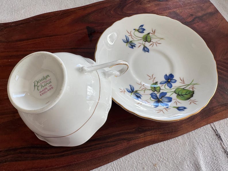 May include: A white teacup and saucer set with a floral design featuring blue flowers and green leaves. The teacup has a handle and the saucer has a gold rim. The teacup has a label on the bottom that reads "Roslyn China Made in England".
