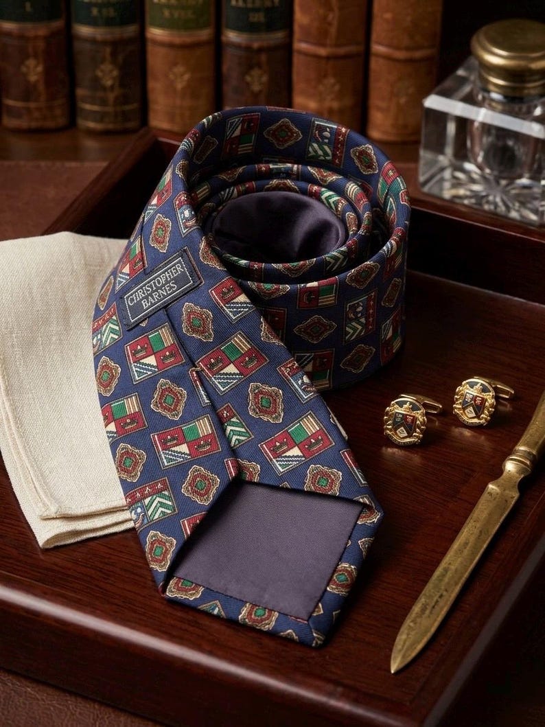 May include: A navy blue necktie featuring a repeating pattern of colourful heraldic crests and geometric shapes. The tie includes a label reading Christopher Barnes. Beside it are two gold-toned cufflinks with crest designs and a metal letter opener.