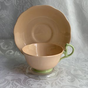 May include: A peach-colored teacup and matching saucer set. The teacup features a light green handle and a light green base, while the saucer has a scalloped edge. Both pieces have a smooth, glossy finish and a consistent solid peach color throughout.