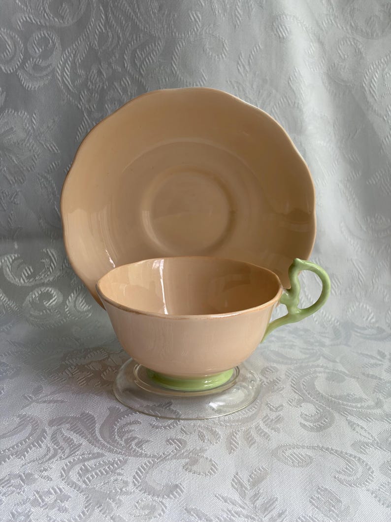 May include: A peach-colored teacup and matching saucer set. The teacup features a light green handle and a light green base, while the saucer has a scalloped edge. Both pieces have a smooth, glossy finish and a consistent solid peach color throughout.