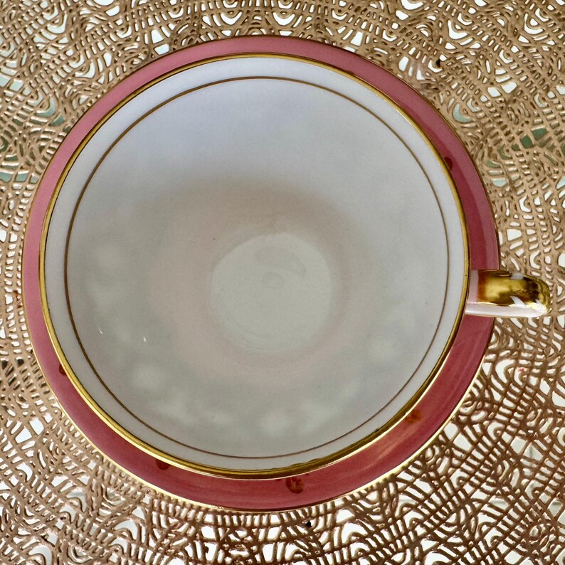 May include: Overhead view of a vintage teacup and saucer set. The cup and saucer are a soft pink with gold trim. The inside of the cup is white. The handle of the cup is gold. The set is on a textured, golden-brown surface.
