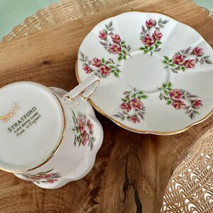 May include: A white teacup and saucer set with a floral pattern. The saucer has a gold rim and the teacup has a gold band around the top. The teacup has the words "Stratford Fine Bone China Made in England" on the bottom.