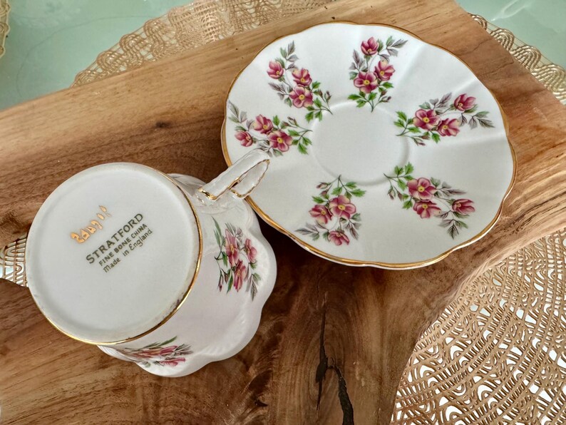 May include: A white teacup and saucer set with a floral pattern. The saucer has a gold rim and the teacup has a gold band around the top. The teacup has the words "Stratford Fine Bone China Made in England" on the bottom.
