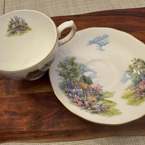 May include: A white teacup and saucer set with a gold trim. The saucer features a cottage scene with a thatched roof, flowers, and trees. The teacup has a floral design.